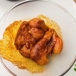 Raw chicken breasts coated in a reddish-brown marinade sit in a clear glass bowl on a white surface.