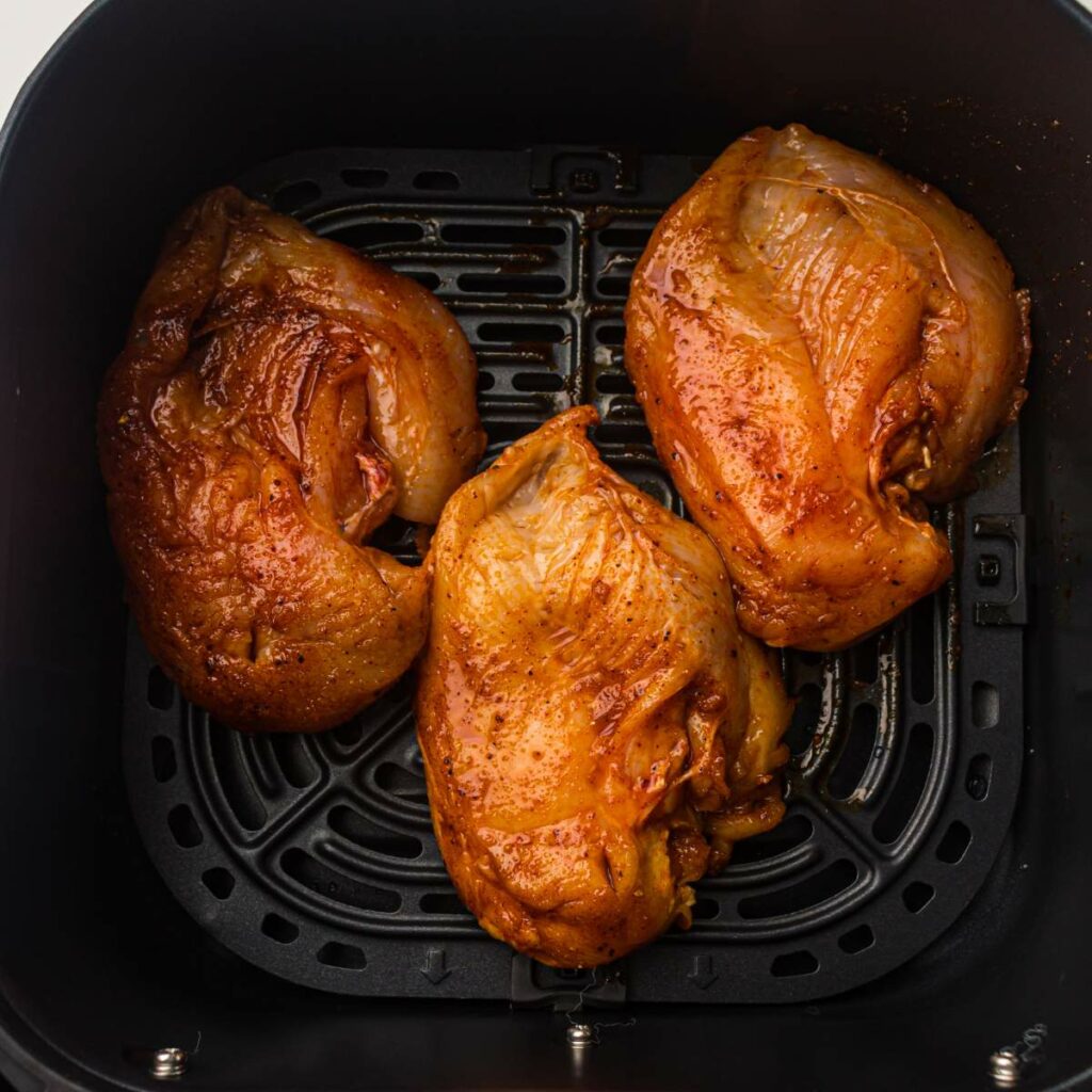 Three seasoned raw chicken breasts are placed inside an air fryer basket, ready to be cooked.