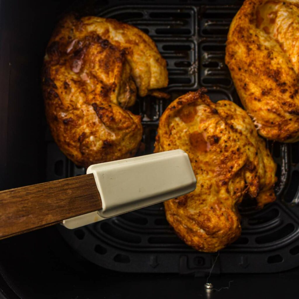 Three pieces of cooked, seasoned chicken breast sit in an air fryer basket with tongs holding one piece.