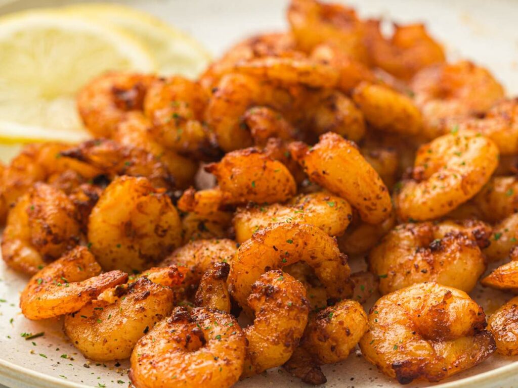 A plate of seasoned, cooked shrimp with two lemon slices in the background.