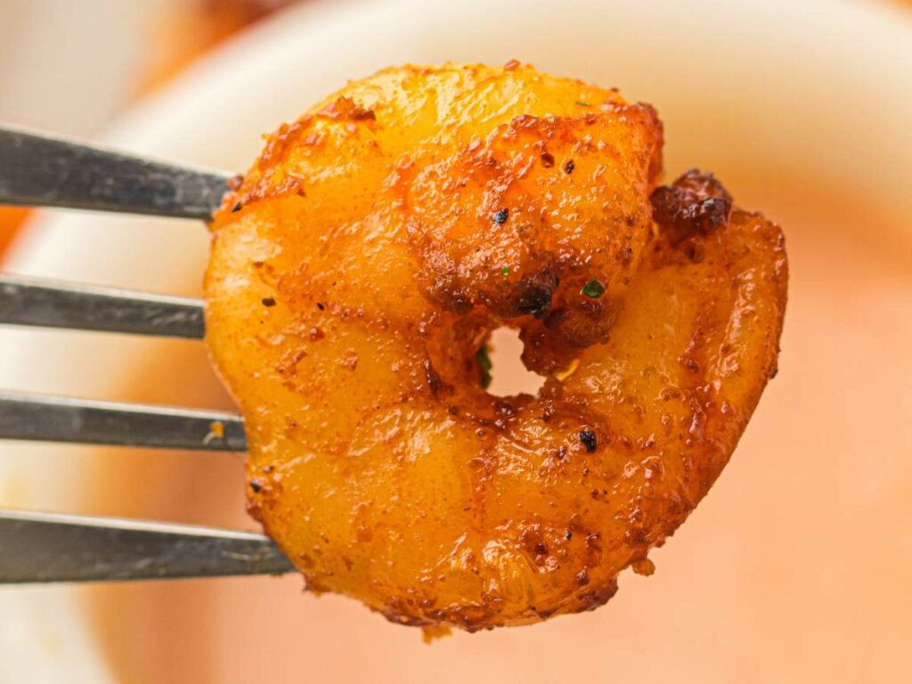 A cooked, seasoned shrimp held by a fork above a bowl of dipping sauce.