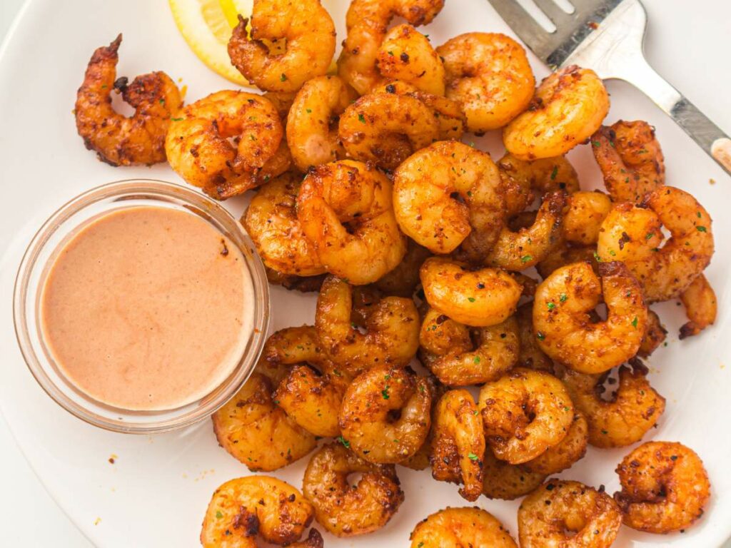 A plate of cooked shrimp served with a small jar of creamy dipping sauce.