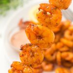 A close-up of a skewer shrimp, with more shrimp and a dipping sauce on a plate in the blurred background.