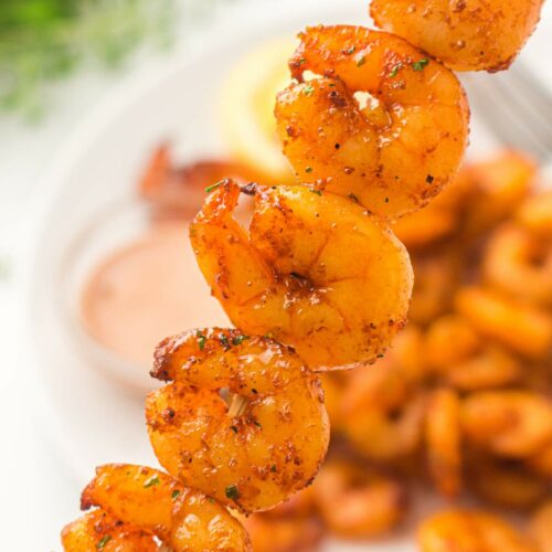 A close-up of a skewer shrimp, with more shrimp and a dipping sauce on a plate in the blurred background.