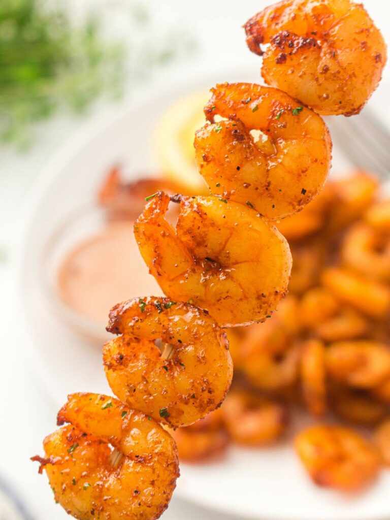 A close-up of a skewer shrimp, with more shrimp and a dipping sauce on a plate in the blurred background.