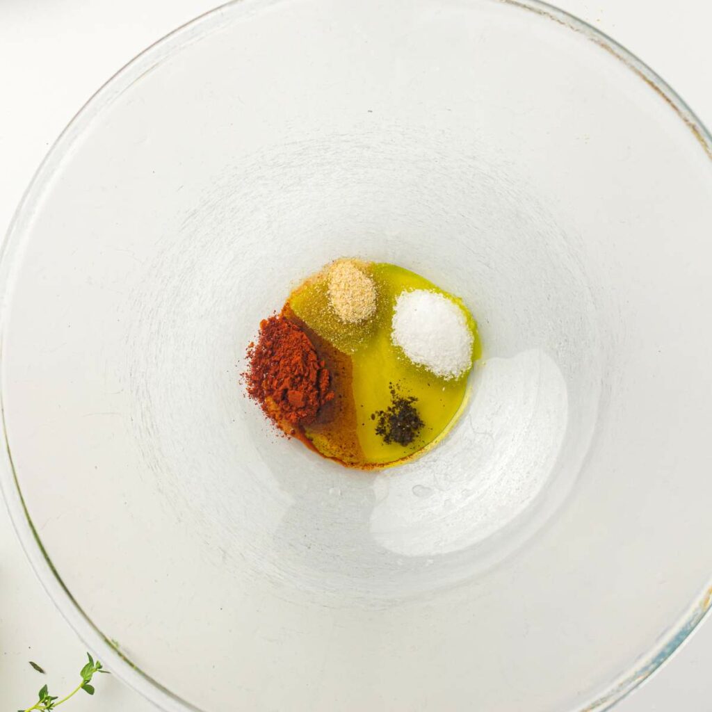 A glass bowl containing avocado oil, paprika, garlic powder, salt, and black pepper arranged separately.