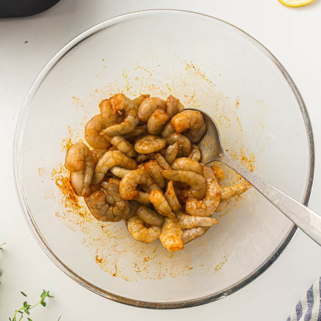 Raw shrimp coated in a seasoning mixture sit in a clear glass bowl with a spoon.