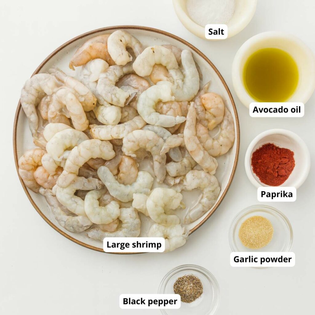 A plate of raw large shrimp surrounded by small bowls containing avocado oil and seasonings.