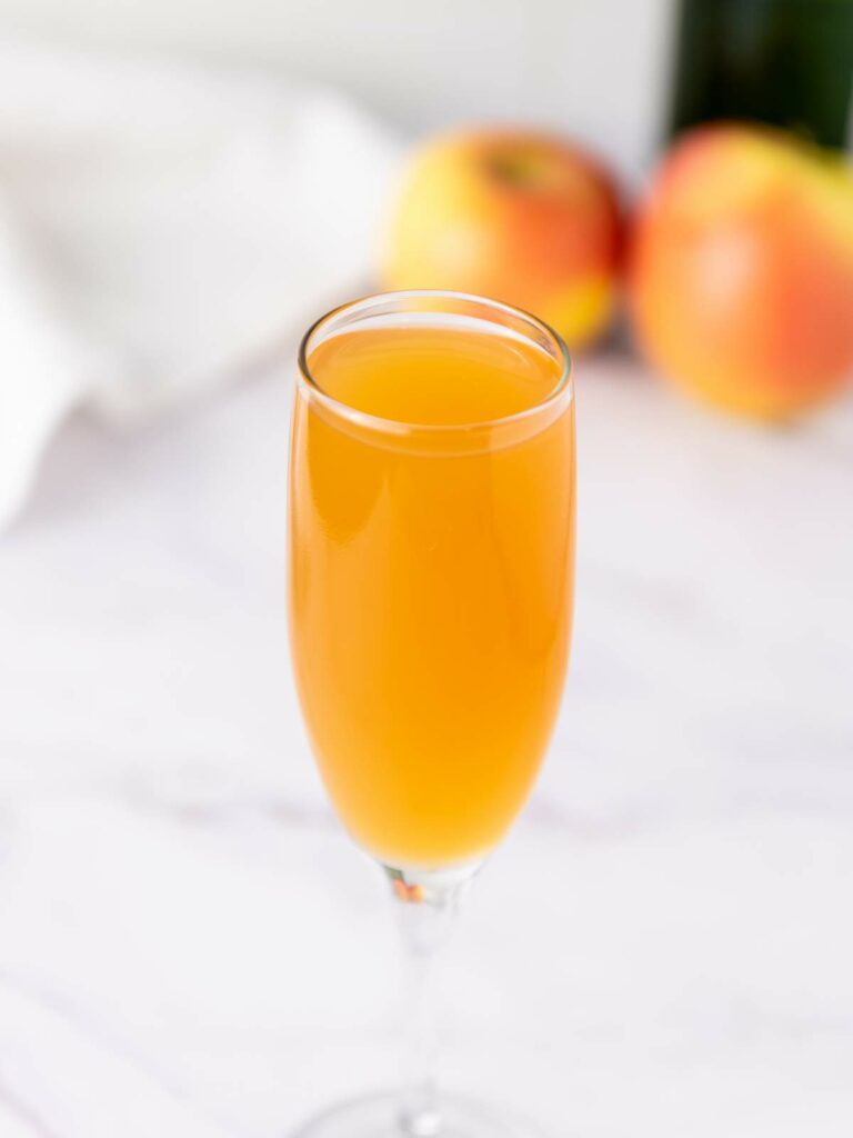 A glass of mimosa in a champagne flute, with two apples and a white cloth in the blurred background.