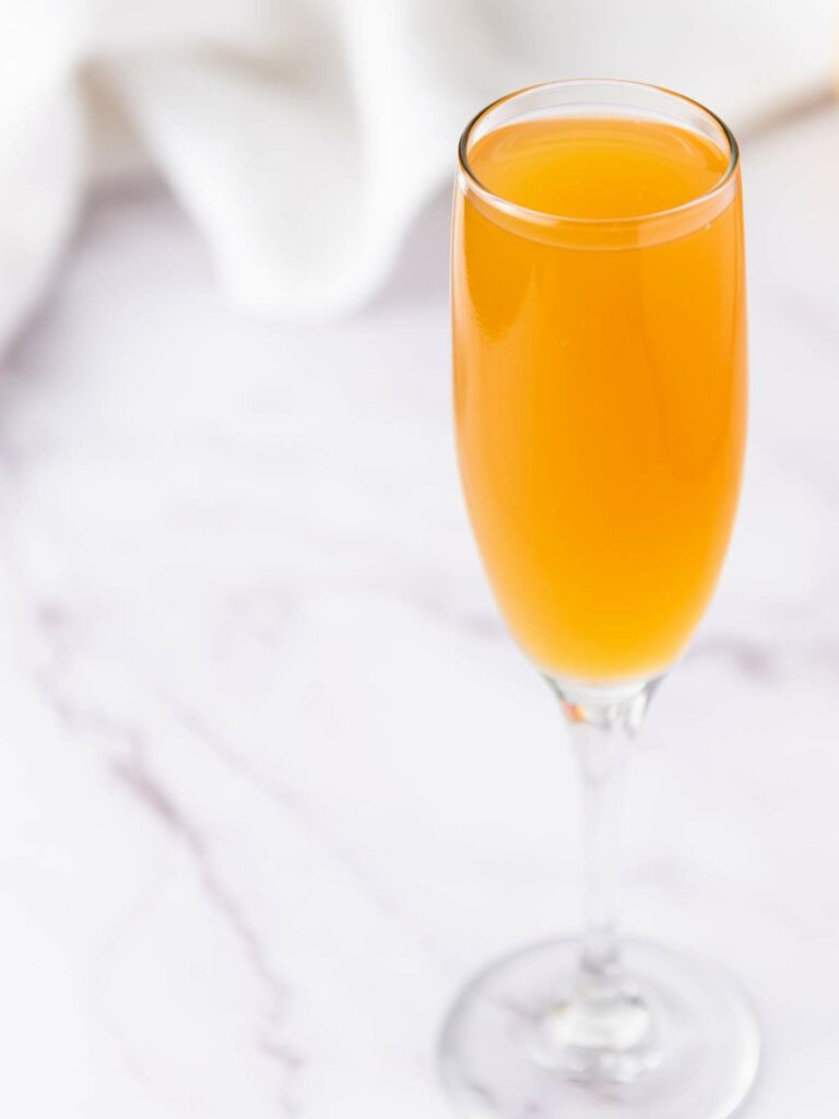 A filled champagne flute containing an orange-colored beverage on a white marble surface.