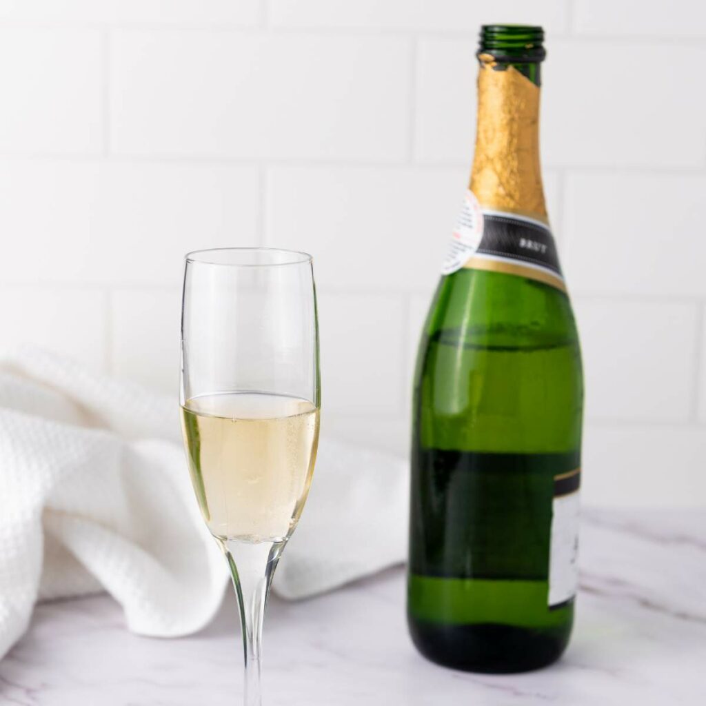 A glass of champagne sits beside an open green champagne bottle on a white marble surface with a white cloth in the background.