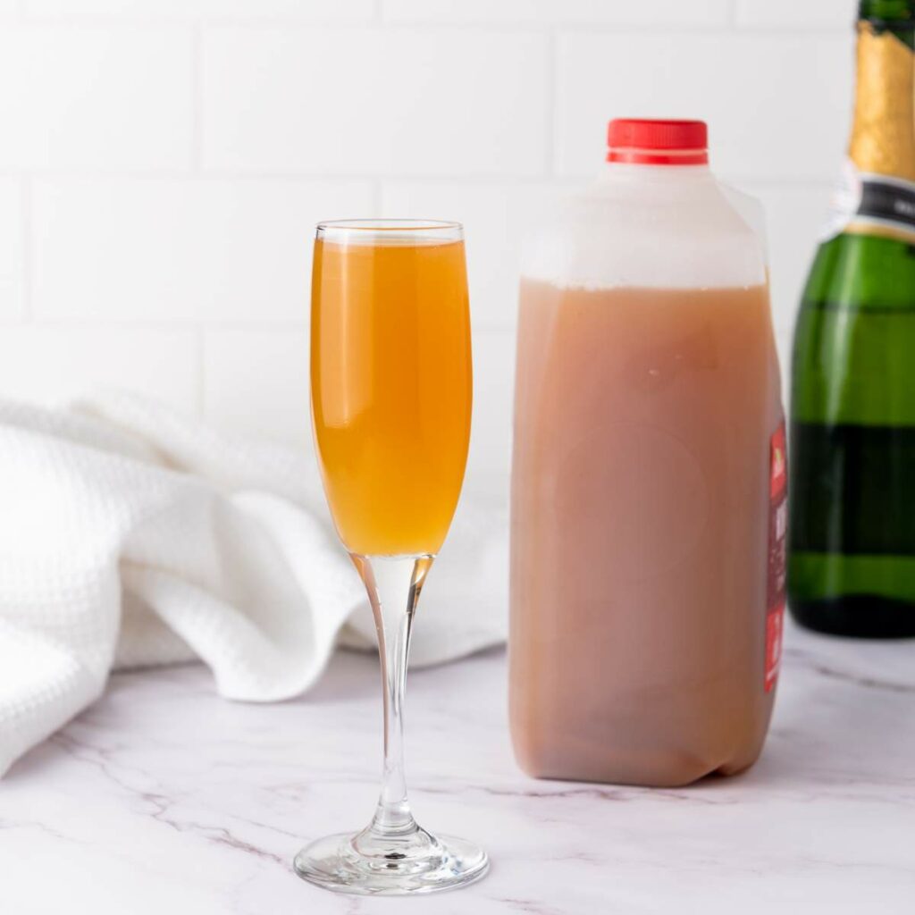 A glass of orange-brown liquid sits in front of a plastic jug of apple cider and a champagne bottle.