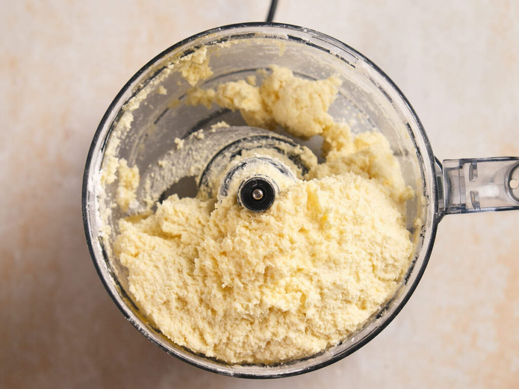 Overhead view of a food processor containing a pale, crumbly mixture.