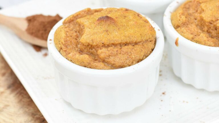 A baked keto soufflé in a white ramekin with a wooden spoon and ground cinnamon in the background.