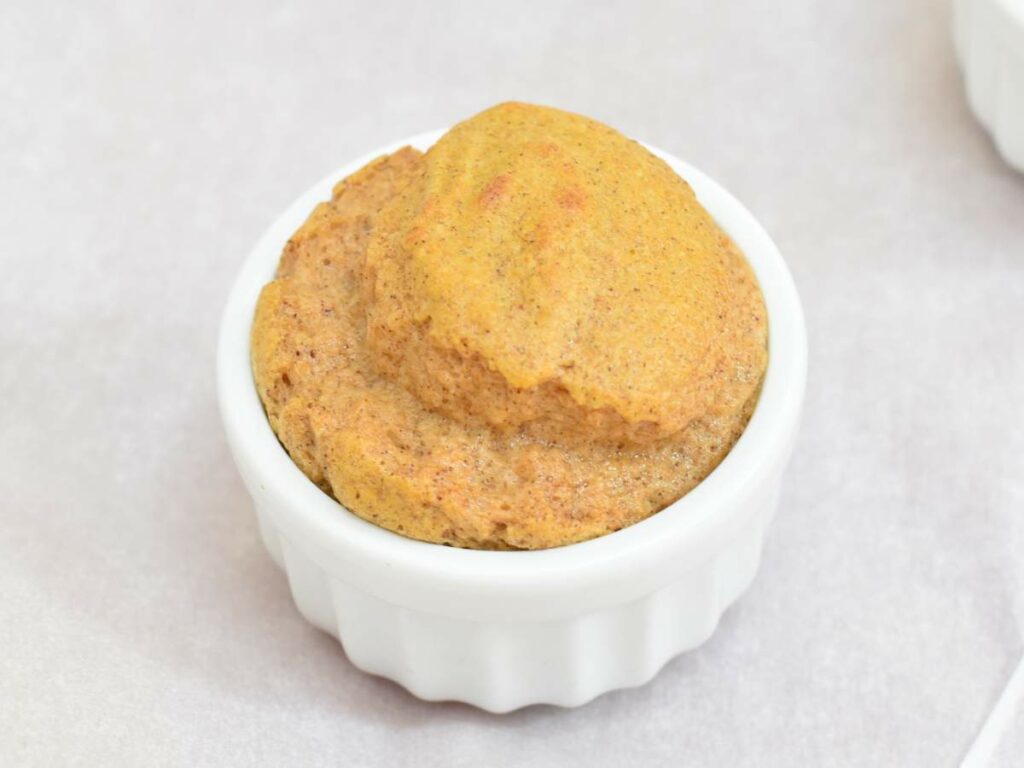 A small white ramekin filled with light brown souffle, set on a plain surface.