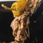 Tongs holding shredded beef and a yellow pepper over a slow cooker.