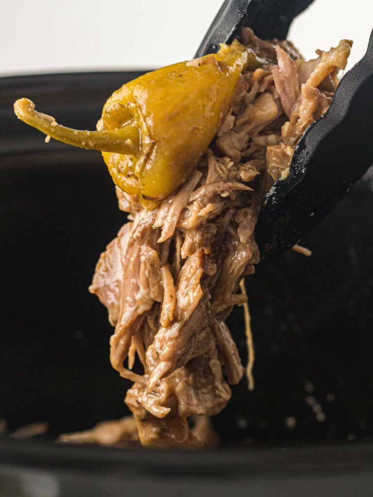 Tongs holding shredded beef and a yellow pepper over a slow cooker.