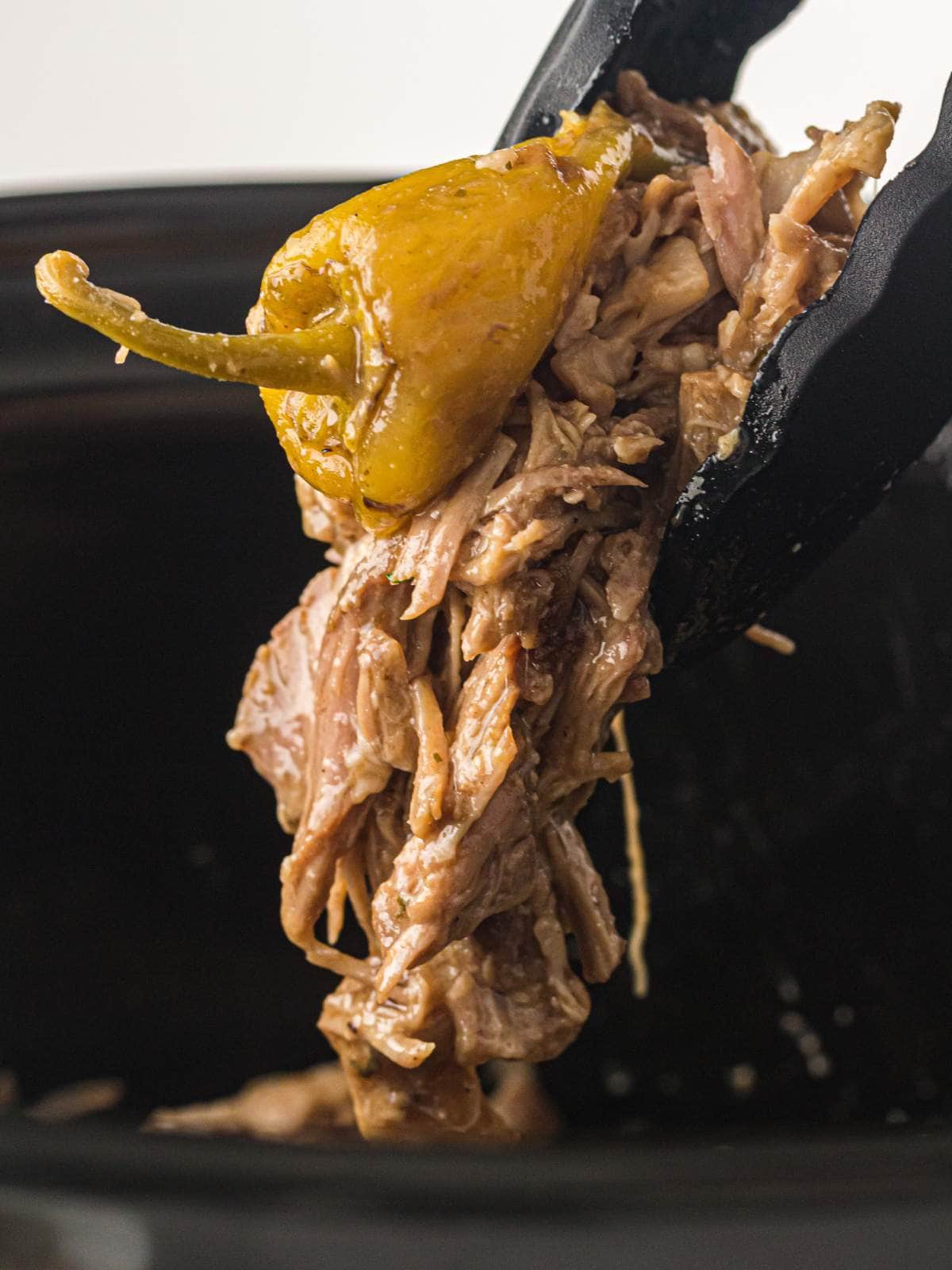Tongs holding shredded beef and a yellow pepper over a slow cooker.