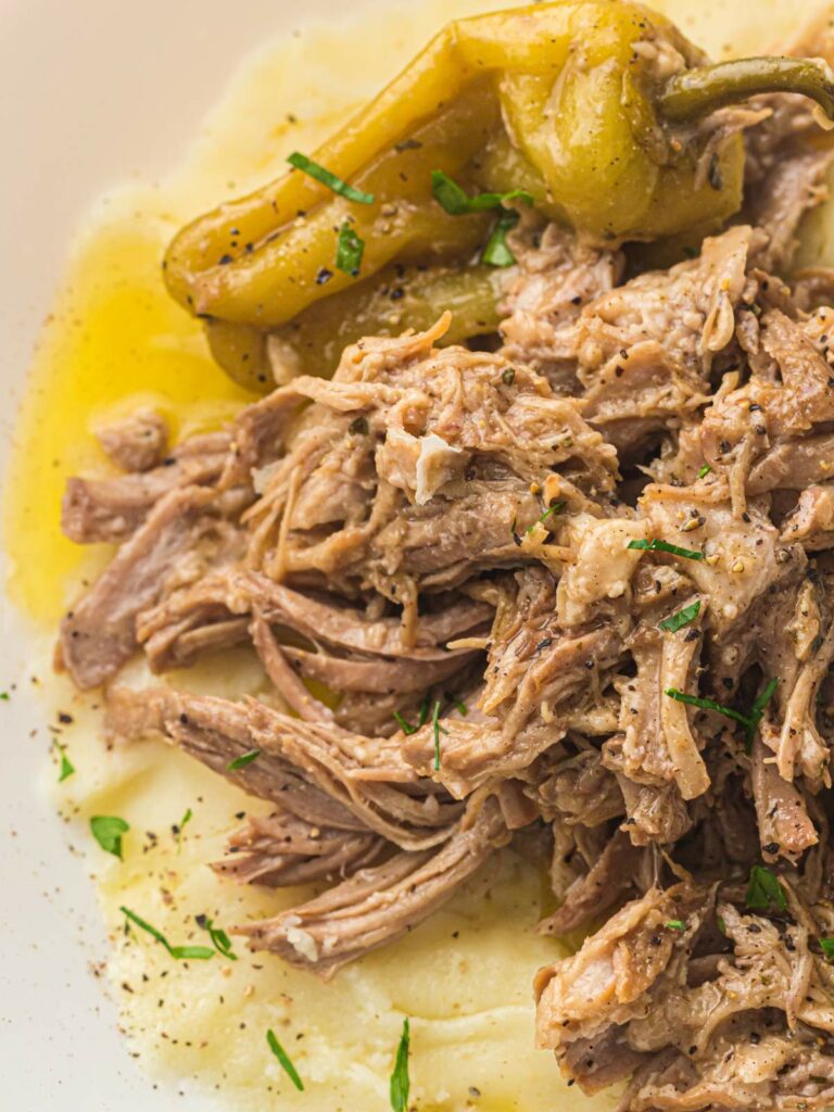 Shredded cooked meat served over mashed potatoes, topped with black pepper, herbs, and a whole pepperoncini pepper.