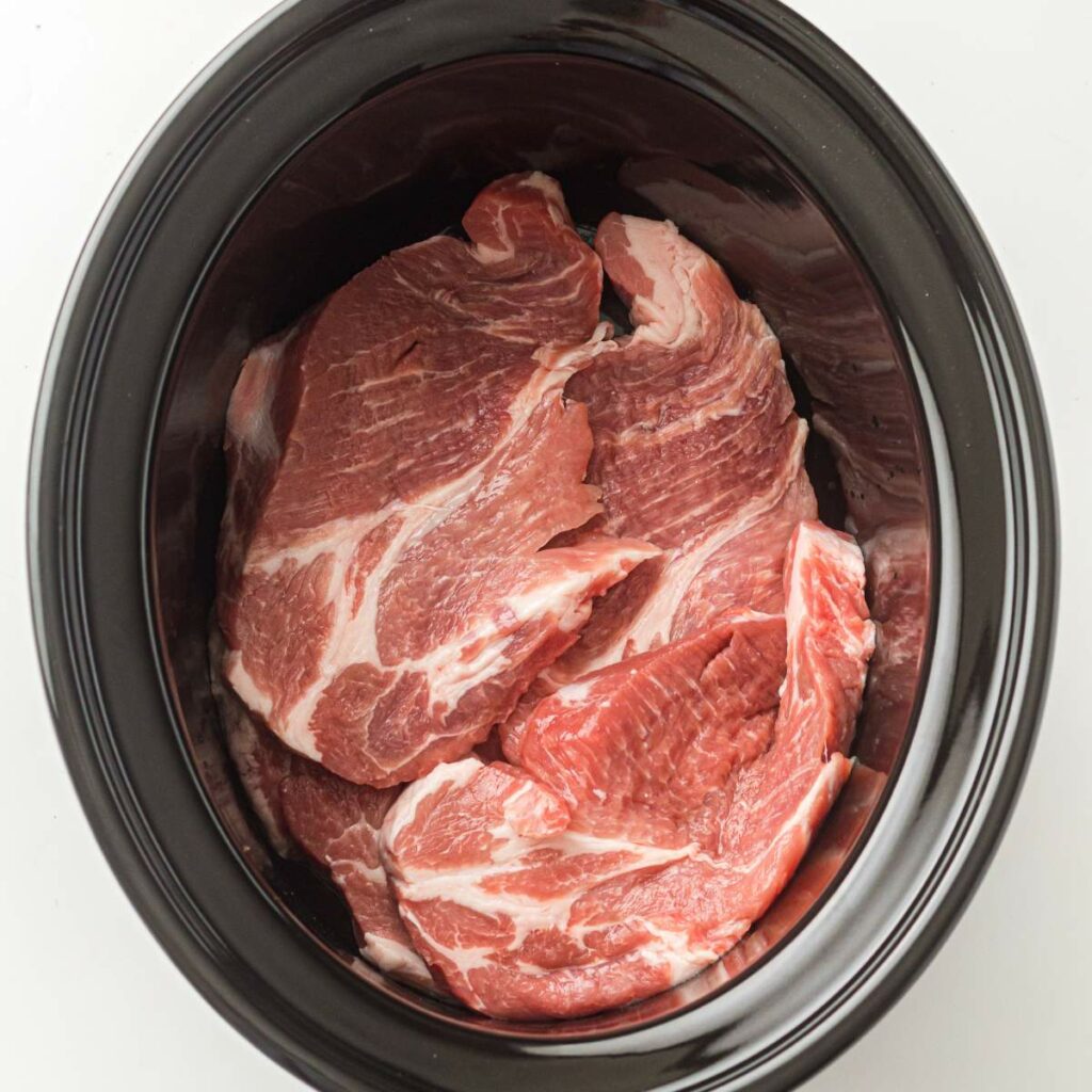 Raw meat pieces placed inside a black slow cooker, ready for cooking.