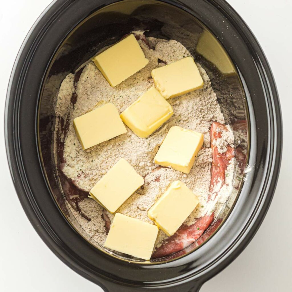 A slow cooker containing raw meat, a layer of powdered seasoning mix, and several slices of butter on top.