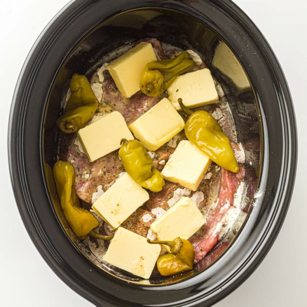 A slow cooker with raw meat, several pepperoncini peppers, and multiple slices of butter arranged on top.