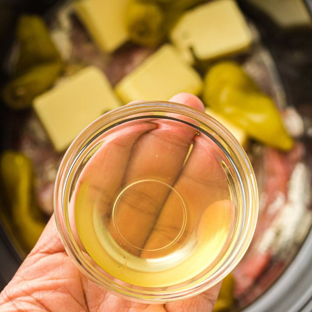 A hand holds a small glass bowl of brine above a slow cooker containing butter, peppers, and other ingredients.