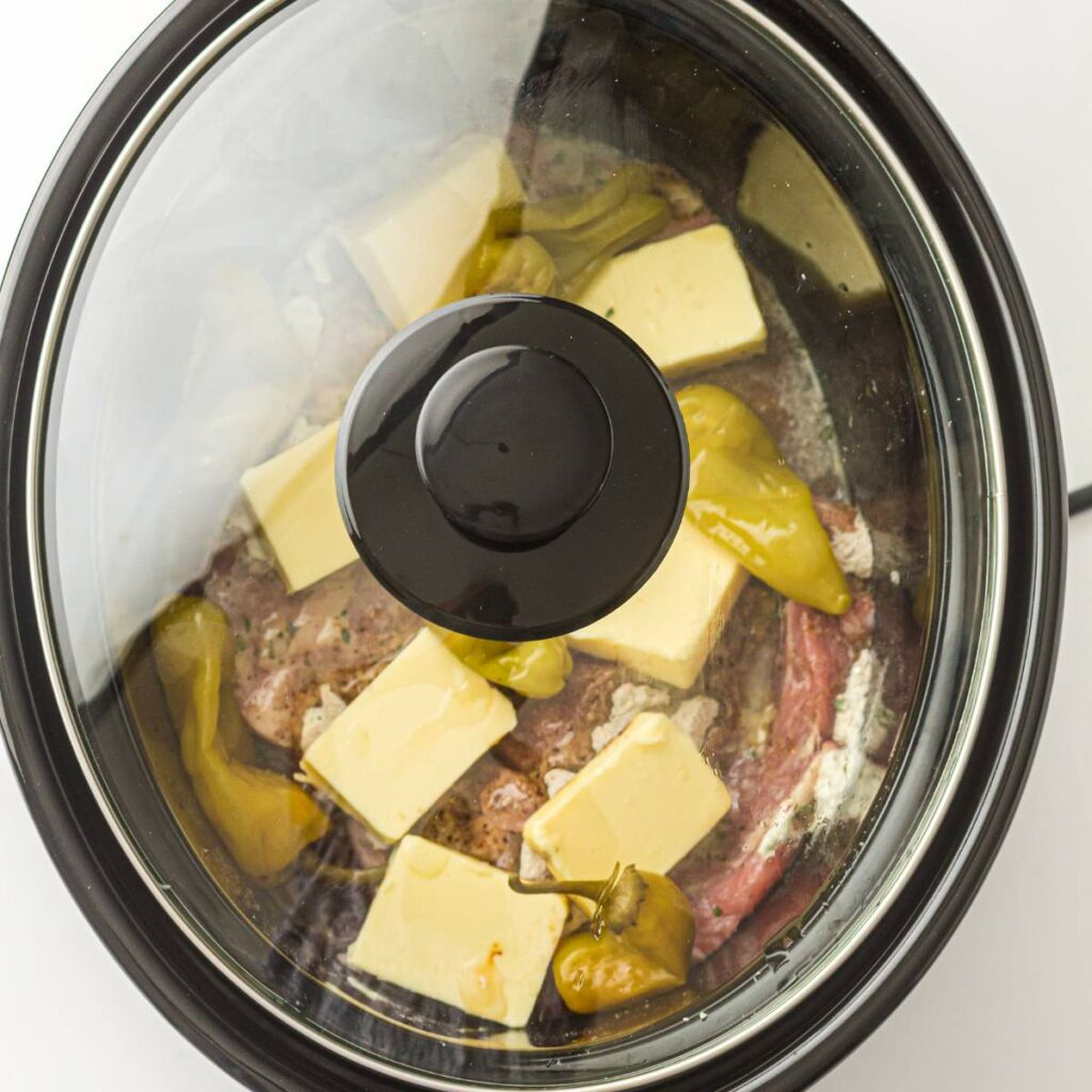 A slow cooker with a glass lid contains uncooked beef, several butter slices, and whole pepperoncini peppers.