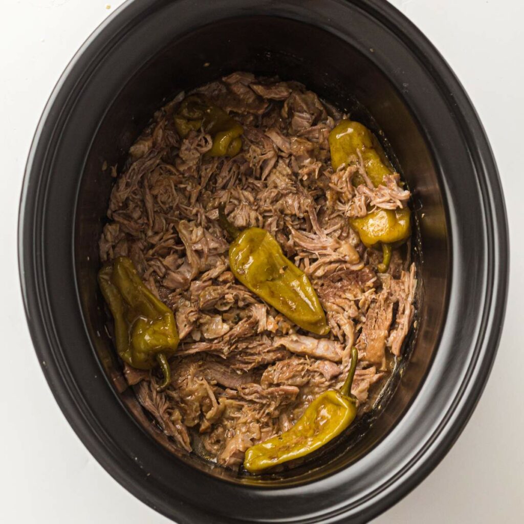 Shredded beef with several whole pepperoncini peppers in a black slow cooker.