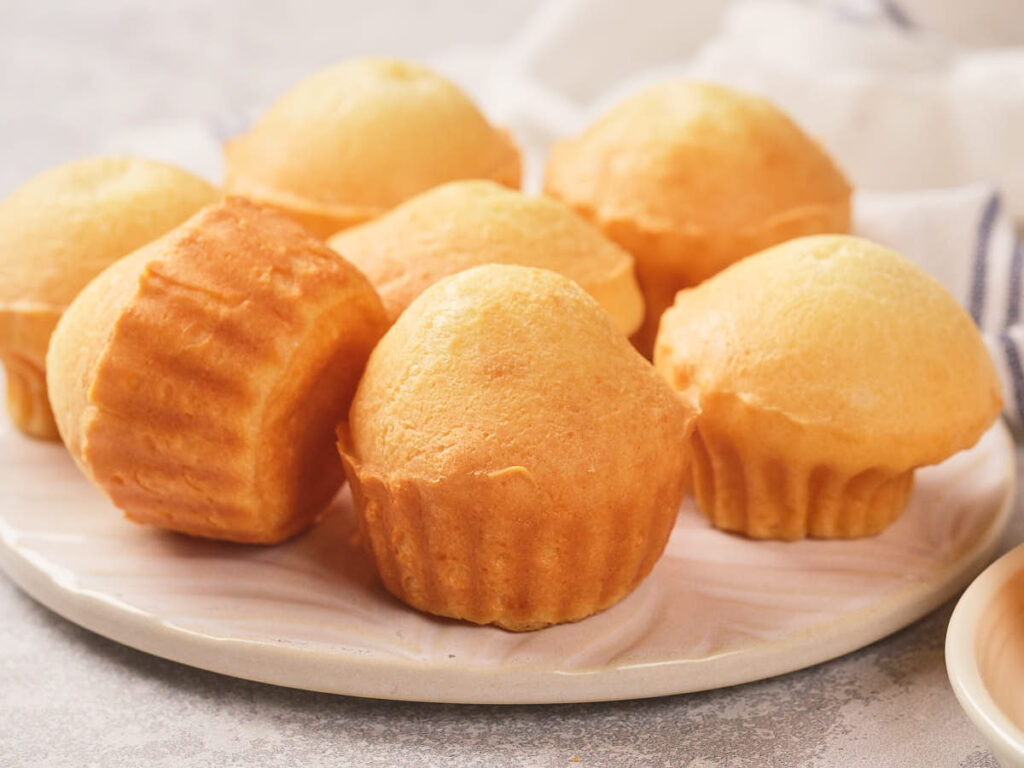 A plate with seven golden brown, plain muffins arranged closely together on a light-colored surface.