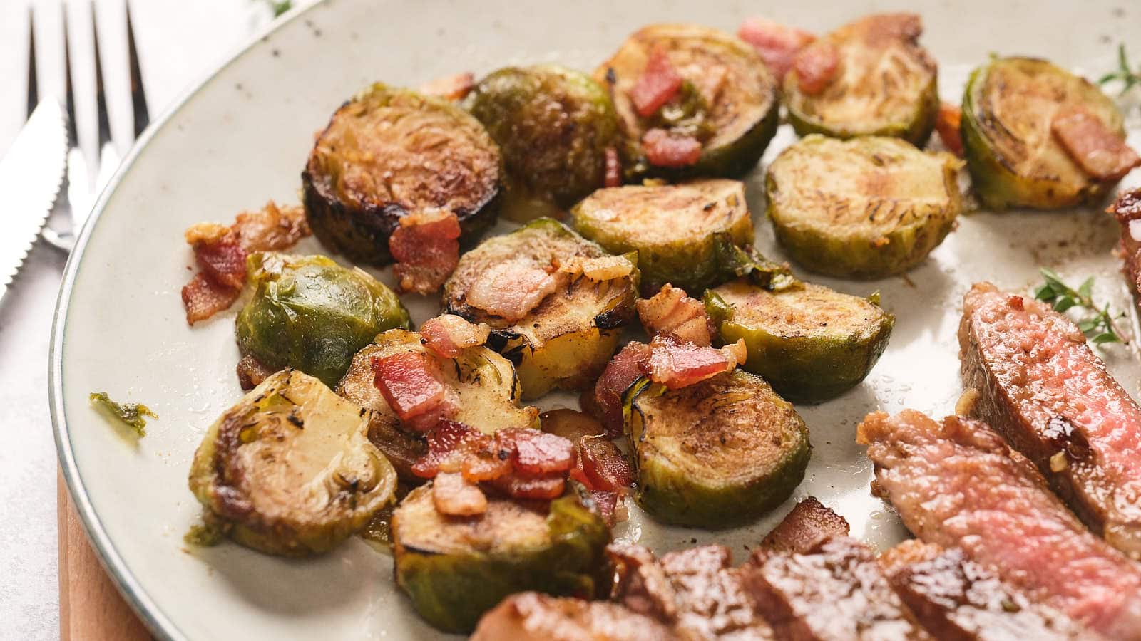 Roasted brussels sprouts with pieces of bacon on a plate next to slices of cooked steak.