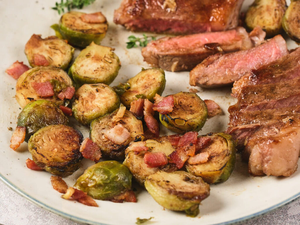 Plate with sliced steak and roasted Brussels sprouts mixed with small pieces of bacon.