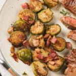 A plate with roasted Brussels sprouts, pieces of cooked bacon, and slices of steak.