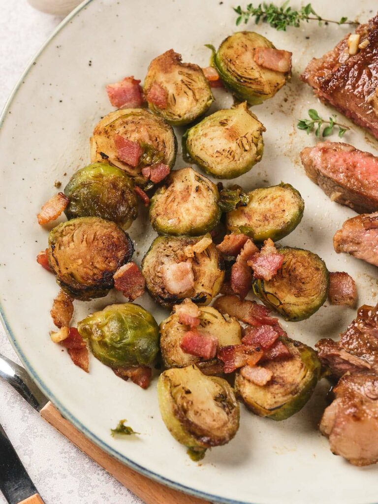 A plate with roasted Brussels sprouts, pieces of cooked bacon, and slices of steak.