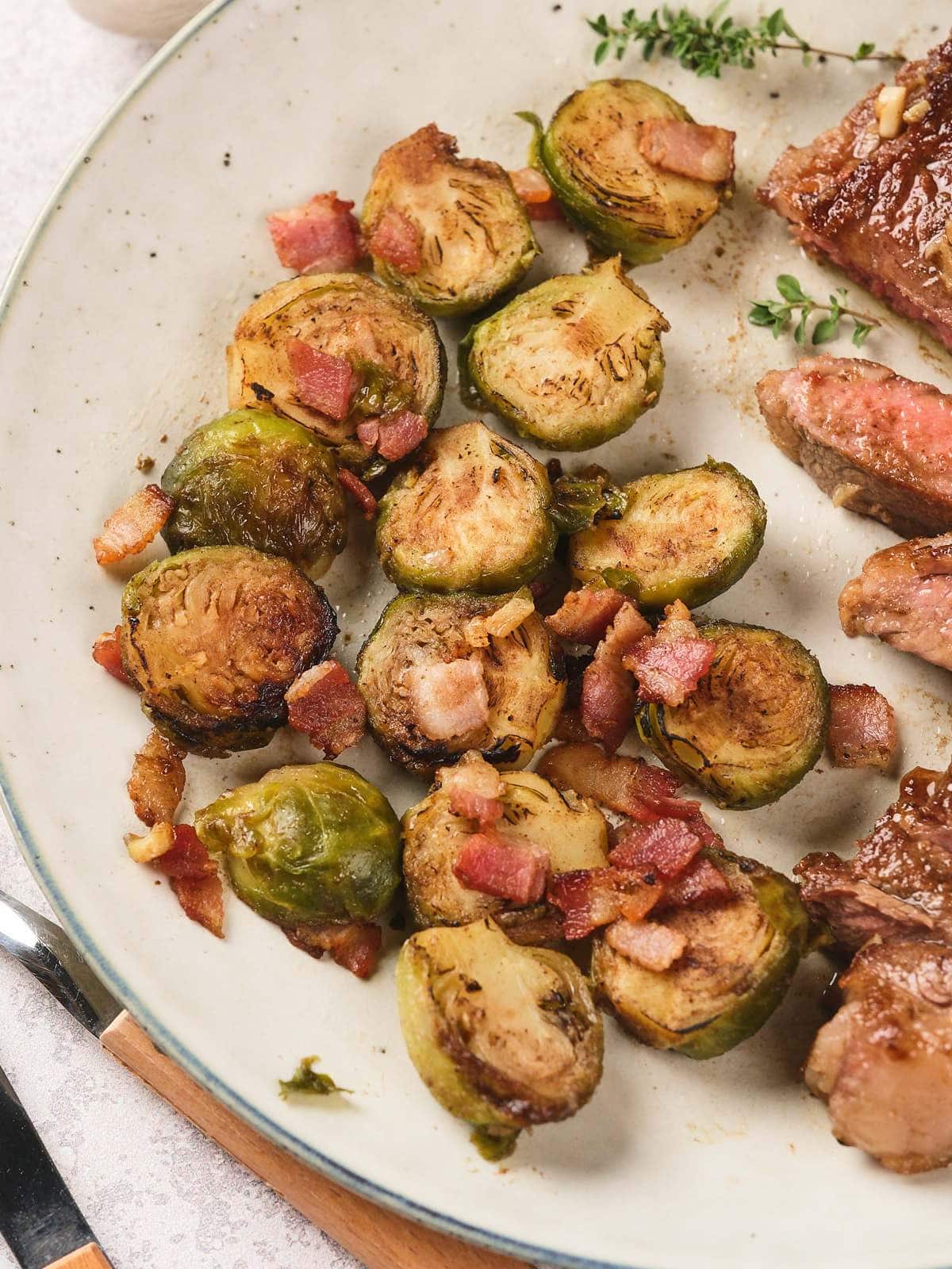 A plate with roasted Brussels sprouts, pieces of cooked bacon, and slices of steak.