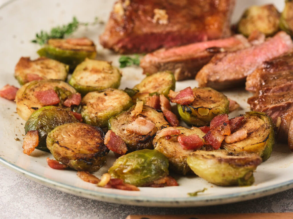 Sliced Brussels sprouts cooked with pieces of bacon, served on a plate next to slices of medium-rare steak.