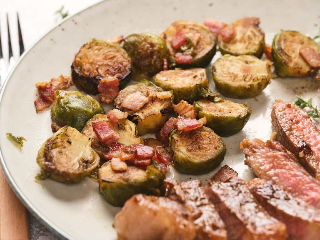 Sliced brussels sprouts and pieces of bacon on a plate next to sliced steak.