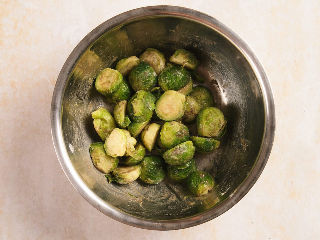 A metal bowl filled with halved Brussels sprouts coated in seasoning on a light-colored surface.