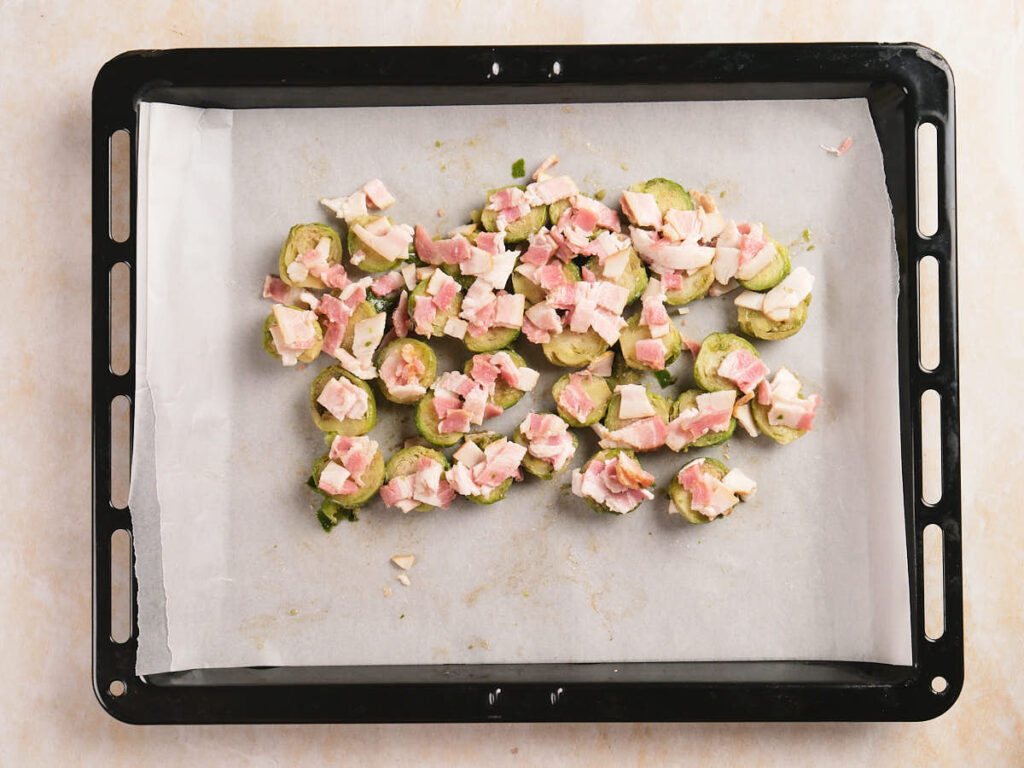 Sliced Brussels sprouts topped with diced ham are arranged on a parchment-lined baking tray.