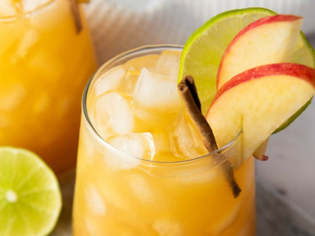 A glass of apple cider mocktail garnished with apple slices, a lime wheel, and a cinnamon stick, with ice cubes visible.