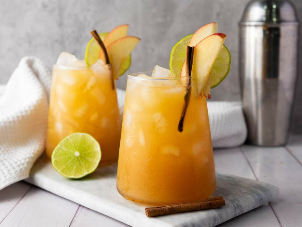Two glasses filled of apple cider mocktail, garnished with apple slices, lime wheels, and cinnamon sticks, placed on a marble board.