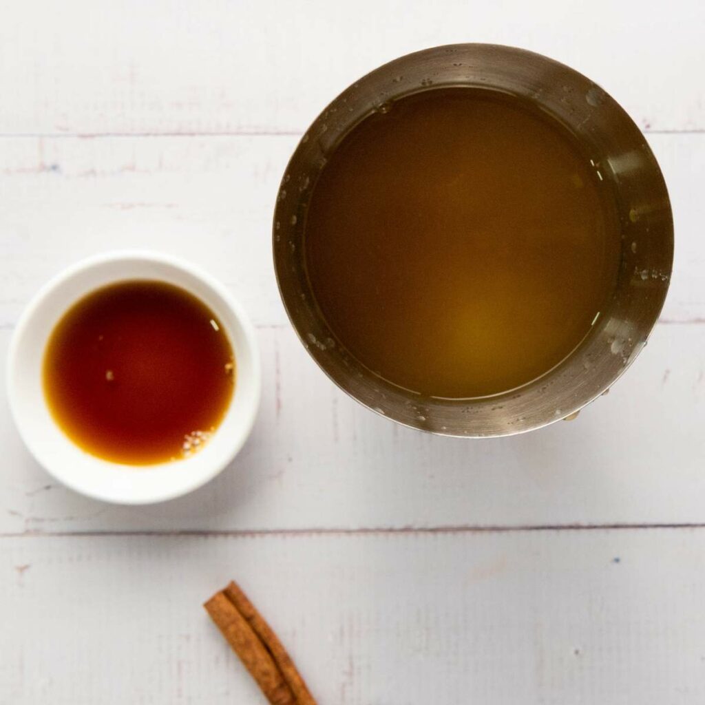 A metal cup filled with brown liquid, a small white bowl containing a darker liquid, and a stick of cinnamon on a white surface.