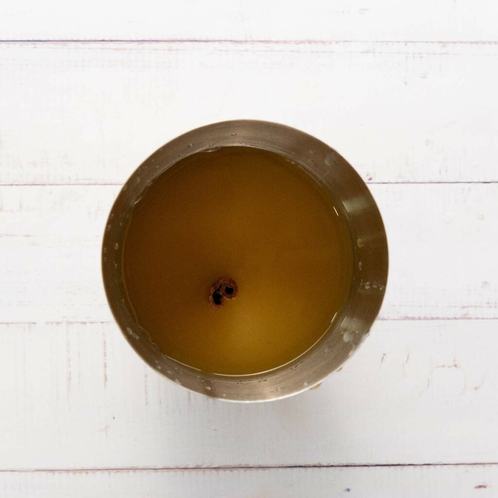 A metal cup filled with yellowish-brown liquid and a single whole spice floating in the center, placed on a white wooden surface.