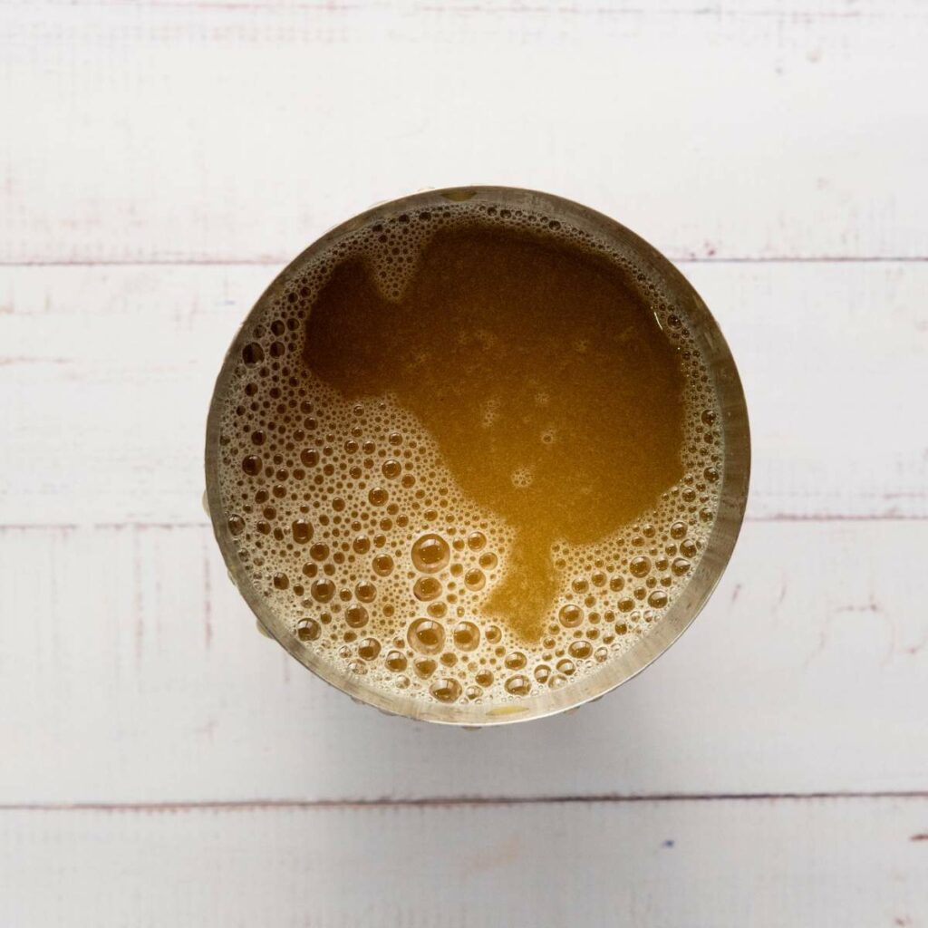 A metal cup filled with frothy brown liquid, viewed from above on a white wooden surface.