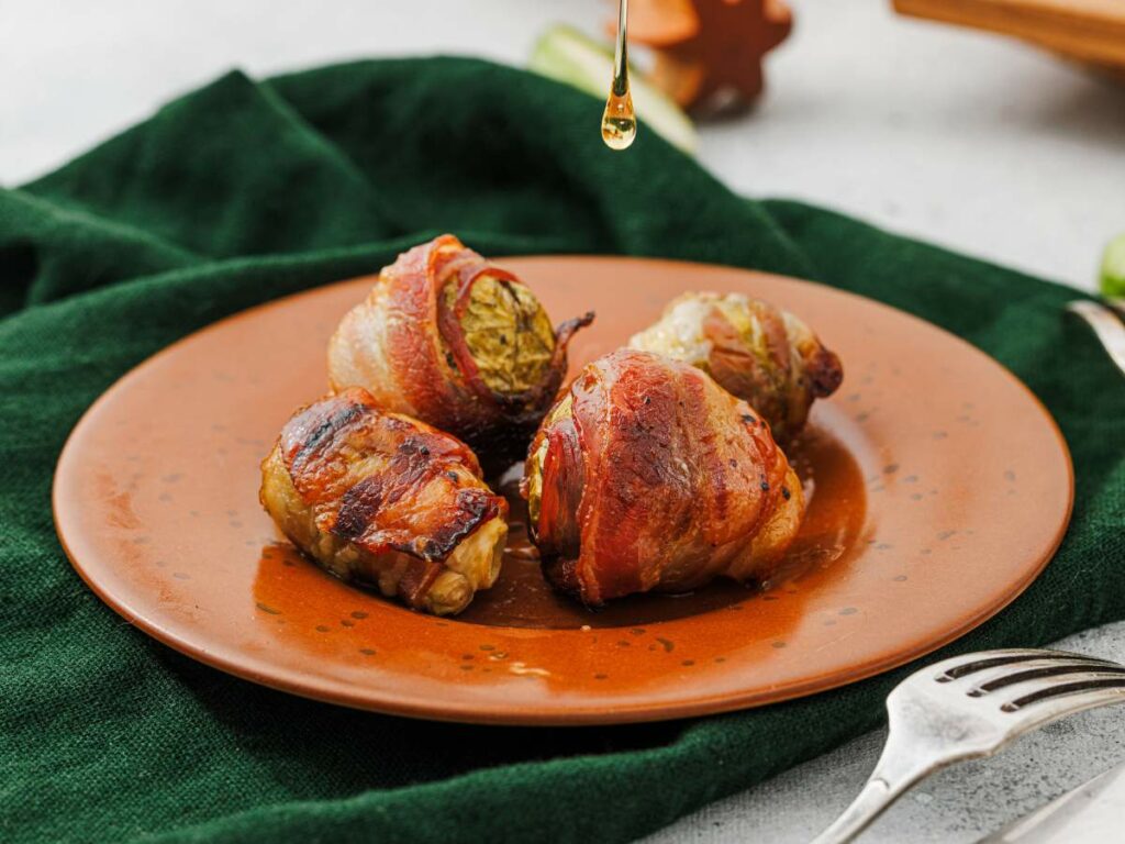 Four bacon-wrapped Brussels sprouts sit on a brown plate, with a drizzle of liquid falling onto them.