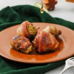 Four bacon-wrapped Brussels sprouts sit on a brown plate, with a drizzle of liquid falling onto them.