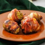 Four bacon-wrapped brussels sprouts on a brown plate with honey being drizzled over them, set on a green cloth.