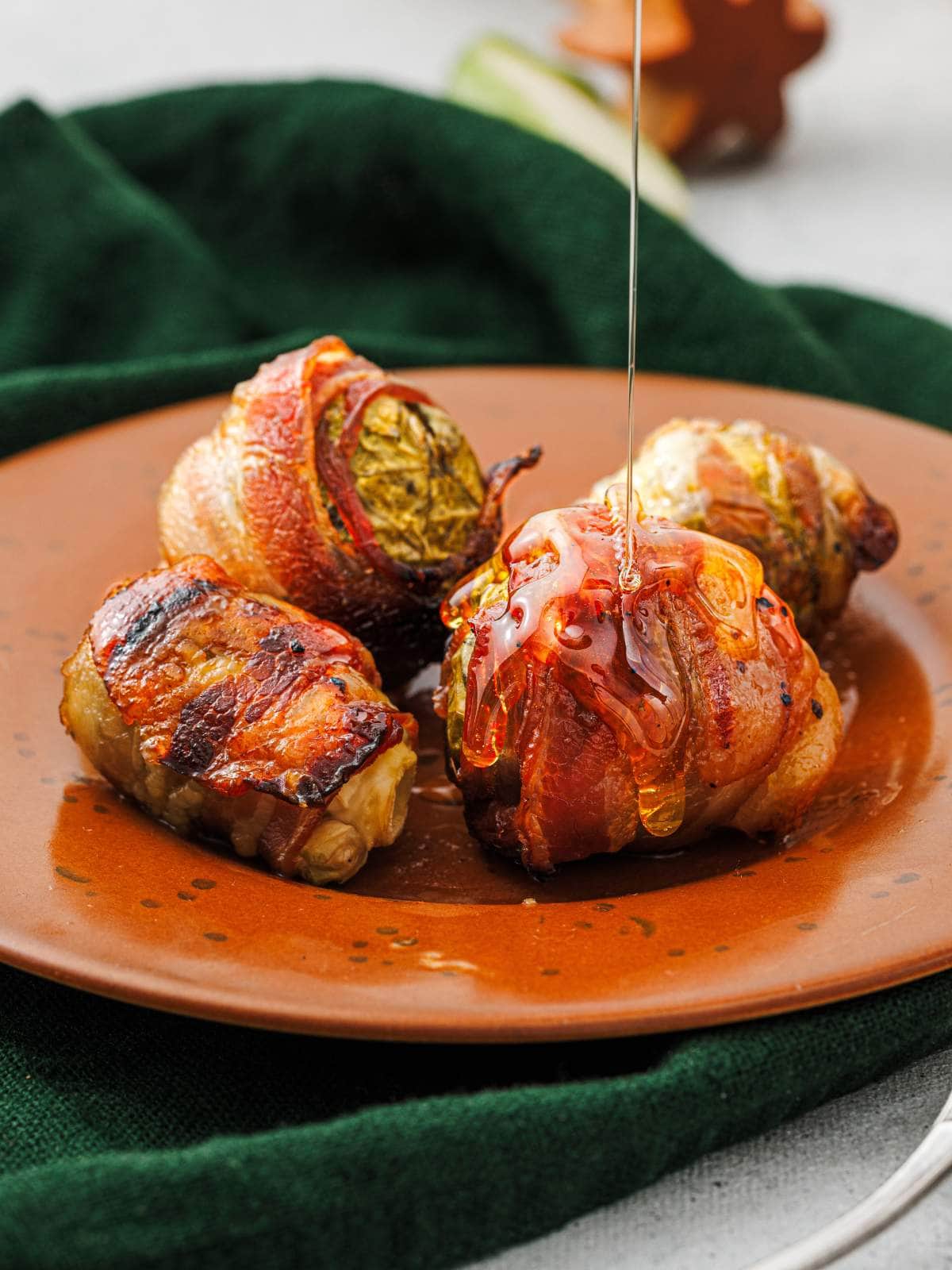 Four bacon-wrapped brussels sprouts on a brown plate with honey being drizzled over them, set on a green cloth.