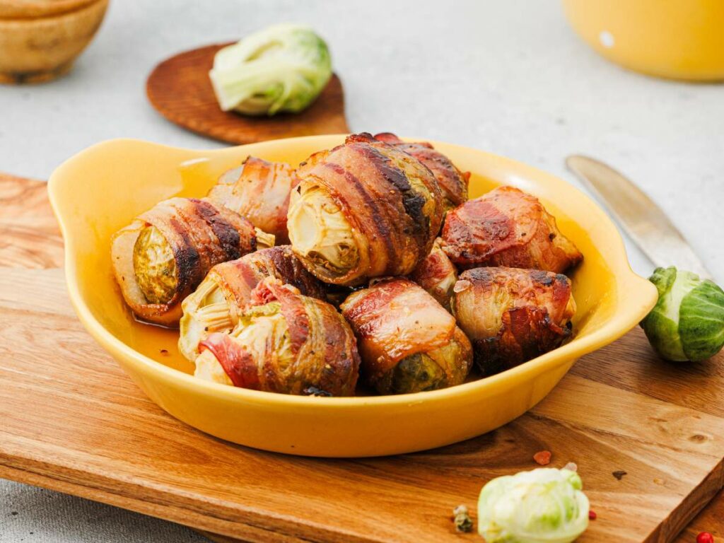 Bacon-wrapped Brussels sprouts are arranged in a yellow dish on a wooden board, with a knife and extra Brussels sprouts nearby.