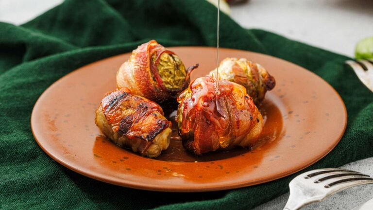 Three bacon-wrapped cabbage rolls on a brown plate, with honey or sauce being drizzled over one, placed on a green cloth with a fork nearby.